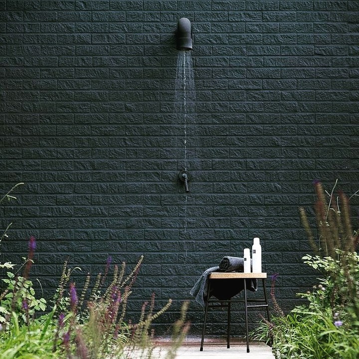 Puštěná černá nerezová sprcha JEE-O soho umístěná na černé stěně, vedle stolička s ručníkem a kosmetikou, v popředí květiny