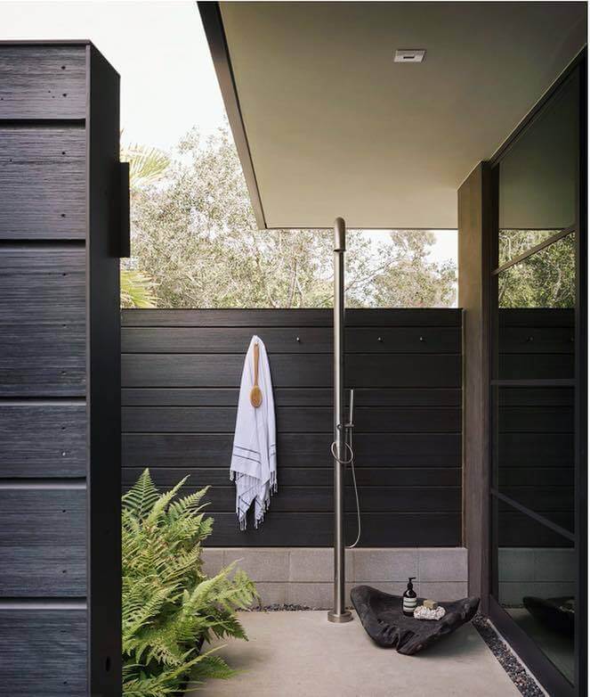 Venkovní sprcha JEE-O original 02 umístěná na terase vedle velkého okna a u vysoké tmavě hnědé dřevěné zdi